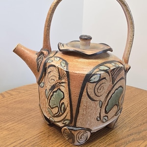 May include: A tan ceramic teapot with a high arched handle and a lid. The body features dark brown hand-painted swirling lines and green leaf-shaped accents. The teapot stands on four small feet and has a short, angled spout on the left side.