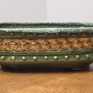 May include: A green ceramic planter with a gold decorative band. The oval-shaped planter has a textured rim and a series of small, raised dots along the base. A decorative piece for plants.