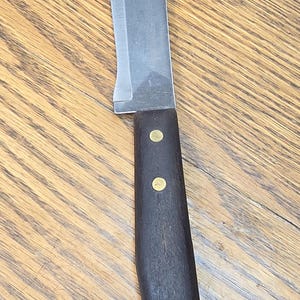 May include: A fixed-blade knife featuring a curved steel blade and a dark brown wooden handle secured with two brass rivets. The handle has a contoured shape for grip, and the blade has a smooth, unpolished finish with a sharp, curved edge.