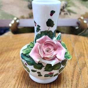 May include: A small, white ceramic vase with a pink rose and green leaves. The vase has a bulbous base and a slender neck. The rose is the focal point, with detailed petals. The vase is decorated with green leaves and red accents.