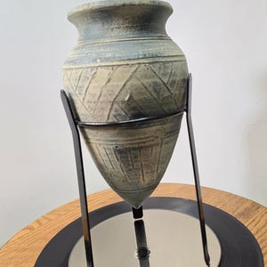 May include: A tapered, pointed-bottom ceramic vessel with a muted blue-green and beige finish, featuring etched geometric patterns around the body. The vessel is supported by a black metal tripod stand that holds it in an upright position.