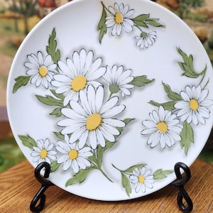 May include: White ceramic plate with a daisy floral design. The plate features large white daisies with yellow centers and green leaves. The plate is displayed on a black metal stand, showcasing a vintage aesthetic.