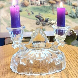 May include: A clear glass candle holder with two lit purple candles. The candle holder has a decorative, scalloped base and two candle cups. The background features a landscape painting.