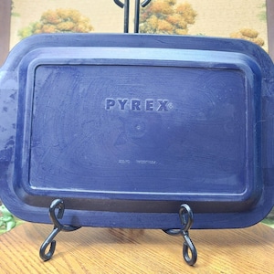 May include: A rectangular, navy blue Pyrex baking dish. The Pyrex logo is centered on the dish. The dish is supported by a black metal stand. The dish has a raised rim and rounded corners.