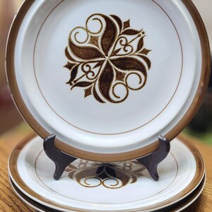 May include: A set of three white ceramic plates with a brown floral design in the center. The plates have a brown rim and are stacked on a wooden surface. The plates are displayed on a wooden plate stand.