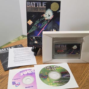 May include: A vintage video game box set for "Battle Sphere" featuring a space battle graphic. Includes the game cartridge, demo CD, and instruction manual. The box is black with gold accents, and the game cartridge is in a white box.