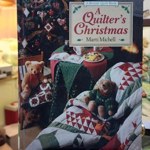 May include: A book cover titled "A Quilter's Christmas" by Marti Michell. The cover features a Christmas tree, a cozy scene with a quilt, and two teddy bears. The book is displayed on a wooden stand.