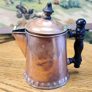 May include: A copper tea kettle with a black wooden handle and lid finial. The kettle has a spout and a hinged lid. The copper has a warm, aged patina. The kettle is likely vintage and would be a decorative item.