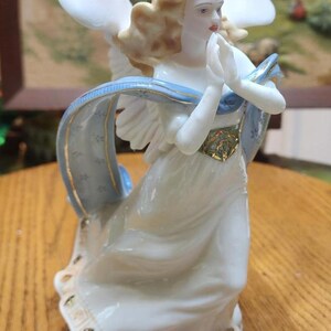 May include: A porcelain angel figurine with flowing white robes, blonde hair, and outstretched wings. A light blue ribbon with gold accents drapes across the figure. The angel's hands are clasped in prayer, and the base has gold detailing.