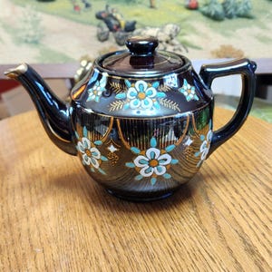 May include: A black ceramic teapot with a lid, spout, and handle. The teapot is decorated with white and turquoise floral designs and gold accents. The teapot is sitting on a wooden surface.