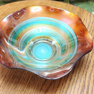 May include: A decorative glass bowl with a unique swirl design. The bowl features a vibrant color palette of turquoise, green, and orange, creating a captivating visual effect. The bowl's rim has a metallic finish, adding a touch of elegance.