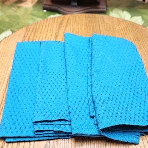 May include: A stack of four folded turquoise napkins with a textured, dotted pattern. The napkins are on a wooden table with a decorative border. A tapestry is visible in the background.