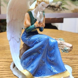 May include: A porcelain angel figurine with a serene expression, seated on a log. The angel wears a blue dress with rose detailing, an orange and green top, and has large white wings. The base is white with a pink rose.