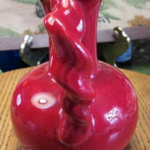 May include: A glossy, red ceramic vase with a bulbous base and a slender neck. The vase features a unique, twisted handle and a smooth, reflective surface. The vase is a decorative item.