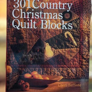 May include: A book titled "301 Country Christmas Quilt Blocks" with a patchwork quilt design on the cover. The cover features a snowman, candy cane, and various quilt block patterns in shades of brown, red, and green. The author's name, Cheri Saffiote, is at the bottom.