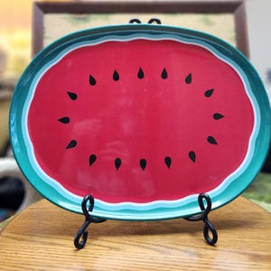 May include: Oval serving tray with a watermelon design. The tray features a red center with black seed details, a white border, and a turquoise outer rim. The tray is displayed on a black metal stand.