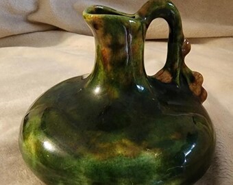 May include: A handcrafted, ceramic pitcher with a rounded body and a curved handle. The pitcher is glazed in shades of green and brown, with a glossy finish. The pitcher is approximately 6 inches tall.