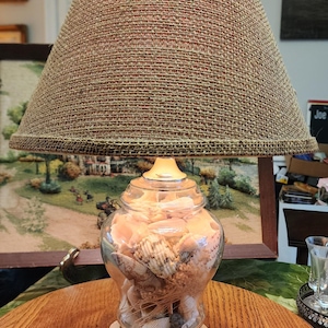 May include: A decorative table lamp with a beige woven lampshade and a clear glass base filled with seashells and sand. The lamp sits on a white base with a seashell design. The lamp is on a wooden table.