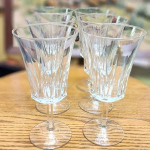 May include: A set of six clear crystal wine glasses. Each glass has a textured, vertical pattern on the bowl and a round base. The glasses are arranged on a wooden surface.