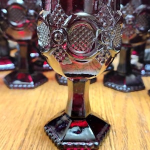 May include: A collection of ruby red goblets with ornate, textured designs. The goblets have a faceted base and a detailed pattern around the bowl. The stem is a solid, dark red color. The goblets are arranged on a wooden surface.