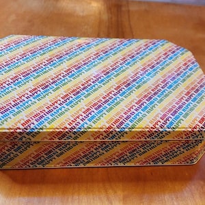 May include: A rectangular metal box with a colorful diagonal stripe pattern. The stripes alternate between yellow, red, blue, and green, with the words "HAPPY BIRTHDAY" printed repeatedly. The box has a shiny, reflective surface.