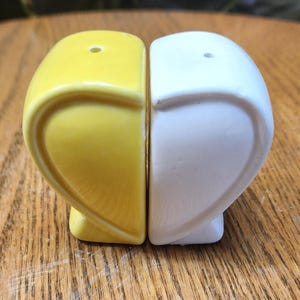 May include: A pair of ceramic salt and pepper shakers. One shaker is yellow, the other is white. Each shaker has a rounded, heart-like shape. The shakers are on a wooden surface.