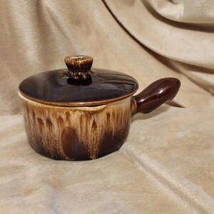 May include: A small, brown ceramic soup pot with a lid and handle. The pot has a dark brown lid and handle, with a lighter brown and tan pattern on the body. The pot is designed for individual servings.