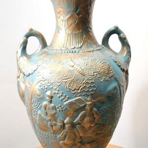 May include: A large, ornate vase with a lid, featuring a teal and gold color scheme. The vase has detailed relief carvings of figures and foliage. It has two handles and stands on a wooden surface.