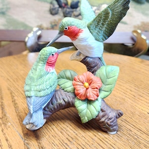 May include: A ceramic figurine of two hummingbirds perched on a brown branch with a red and orange flower. The hummingbirds have green, red, and white coloring. One bird has its wings spread. The figurine is on a wooden table.