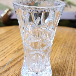 May include: A clear, small glass vase with a starburst pattern. The vase has a flared top and a faceted design, sitting on a wooden surface. The vase is approximately 4 inches tall.