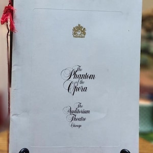 May include: A vintage program for "The Phantom of the Opera" at The Auditorium Theatre in Chicago. The cover is white with gold accents, including a crest and elegant script. A red tassel hangs from the side. The program is displayed on a black metal stand.