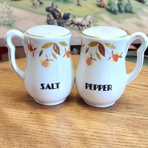 May include: A pair of white ceramic salt and pepper shakers with handles. Each shaker is decorated with a gold rim and a floral pattern in shades of brown, orange, and yellow. The word "SALT" and "PEPPER" are printed in black.