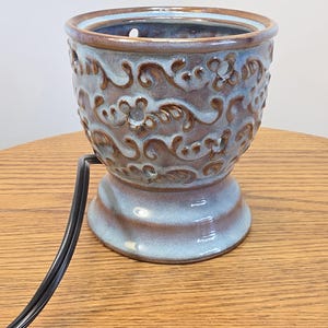 May include: A ceramic wax warmer featuring a blue and brown glazed finish with a raised floral and scroll pattern. The cylindrical vessel has a wide rim, a flared base, and a black electrical cord extending from the side for power connection.