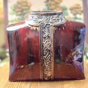 May include: A decorative ceramic vase shaped like a jacket, in shades of burgundy and brown. The vase has a high collar and a central panel with floral and leaf designs in gold. The vase is on a wooden surface.