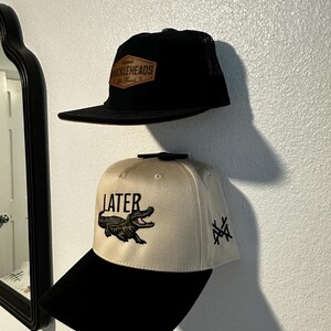 Puede incluir: Se muestran dos gorras de béisbol. La gorra superior es negra con una parte trasera de malla y un parche marrón que dice "BUCKLEHEADS". La gorra inferior es beige con una visera negra, la palabra "LATER" y un cocodrilo.