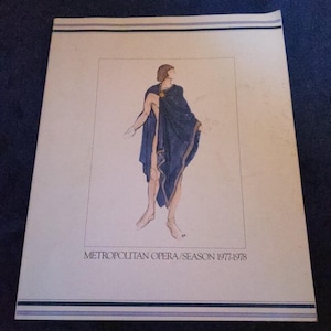 May include: A print featuring a watercolor illustration of a figure draped in a dark blue cloak with gold accents. The figure is standing, and the print includes the text "METROPOLITAN OPERA/SEASON 1977-1978" at the bottom, with blue and black stripes at the top.