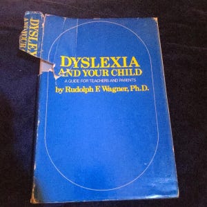 May include: A blue book titled "Dyslexia and Your Child" by Rudolph F. Wagner, Ph.D. The book's cover features the title in large yellow letters, with the subtitle "A Guide for Teachers and Parents." The book is slightly damaged.