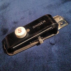 May include: A vintage Singer sewing machine attachment, black with a white knob featuring a red "S". The attachment has a silver metal component and the Singer logo is visible. The item is on a blue surface.