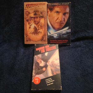 May include: Three VHS tapes featuring Harrison Ford. Titles include "Indiana Jones and the Last Crusade", "Air Force One", and "The Fugitive". The tapes have colorful movie artwork and text.