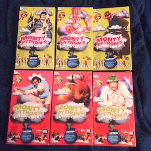 May include: Six VHS tapes of Monty Python's Flying Circus are displayed. The top row features yellow cases with illustrations of characters, while the bottom row has red cases. Each tape has the show's logo and episode titles.