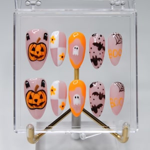 May include: A set of Halloween-themed press-on nails. Designs include orange jack-o'-lanterns, ghosts, bats, and spiderwebs. The nails are displayed in a clear acrylic case with a gold-colored stand.