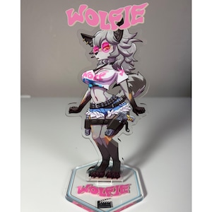 Puede incluir: Figura acrílica transparente de un personaje parecido a un lobo con pelaje gris, que lleva una camisa blanca, una falda azul y gafas de sol. La figura tiene una pancarta rosa "WOLFIE" encima y una base rosa con el nombre. El personaje sostiene un arma.