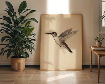 May include: A framed print featuring a detailed black and white illustration of a hummingbird in flight. The artwork is set against a cream-colored background and framed in a light wood. A large potted plant is to the left of the framed print.
