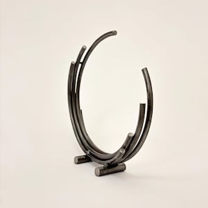 May include: A dark metal sculpture with a circular design. The sculpture is made of curved metal rods and has a minimalist aesthetic. The base consists of two horizontal rods. The sculpture is approximately 10 inches tall.