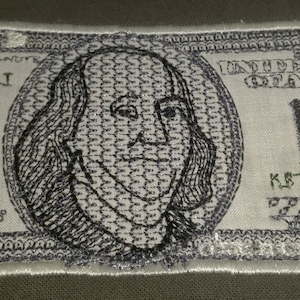 Puede incluir: Un parche bordado de un billete de 100 dólares con un retrato de Benjamin Franklin. El parche es blanco con bordado negro y verde. El número de billete es "KB46279860T".