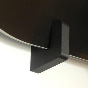 May include: Close-up of a black metal snowboard wall mount. The mount is an L-shape design, with a flat base and a curved edge to hold the snowboard. The snowboard is black with a silver edge.