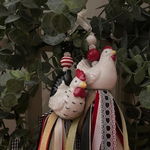 May include: Two ceramic chicken ornaments with red combs and yellow beaks. The chickens are adorned with colorful ribbons and beaded accents in red, white, and black. The ornaments are hanging in front of a green leafy background.