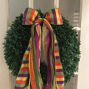 May include: A round green boxwood wreath adorned with a large decorative bow. The bow features long, trailing ribbons in a vibrant plaid pattern of orange, purple, yellow, and green, alongside solid purple, lime green, and orange glittered ribbons.