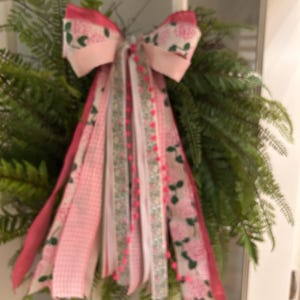 May include: A decorative wreath composed of lush green fern fronds adorned with a large, multi-layered bow. The bow features ribbons in shades of pink, white, and green, including patterns of floral prints, solid pink, and a thin string of small pink pom-poms.