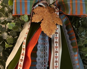 Op de afbeelding: Een decoratieve strik met blauw, groen en oranje geruit lint, effen crèmekleurige en donkergroene linten, en een lichtblauwe kanten rand. Een glinsterend koperkleurig esdoornbladornament is in het midden van de strik geplaatst, die op groen bladwerk ligt.